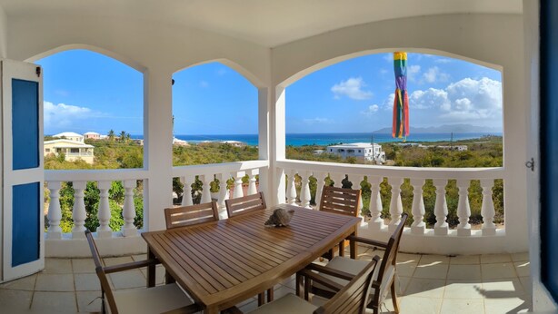 Une terrasse couverte avec une table et des chaises en bois, des balustrades blanches et une vue sur la mer et les bâtiments.