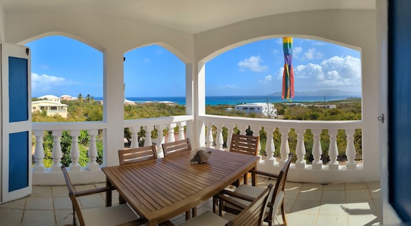 Un porche cubierto con mesa y sillas de madera, balaustradas blancas y vistas al mar y edificios.