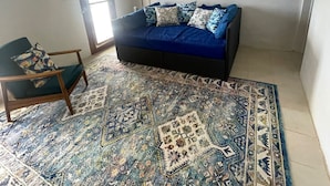 Une pièce avec un canapé bleu, un tapis à motifs, un ventilateur de plafond et une fenêtre avec des stores.