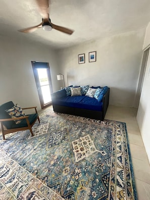 Una habitación con un sofá azul, un tapete estampado, un ventilador de techo y una ventana con persianas.