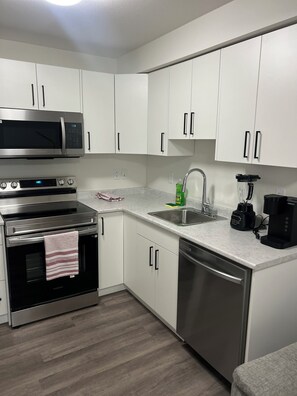 A modern kitchen with white cabinets, stainless steel appliances, a microwave, oven, dishwasher, and a coffee maker. - Warm & Cozy Guest Basement Suite. (Saskatoon)