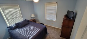 Une chambre à coucher avec un lit, deux fenêtres équipées de stores, une table de chevet avec une lampe, un téléviseur posé sur un meuble en bois et un sol recouvert d’une moquette.