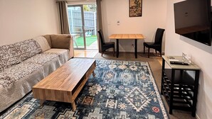 Una sala de estar con sofá, mesa de centro, alfombra estampada, mesa de comedor, sillas y televisor en la pared.