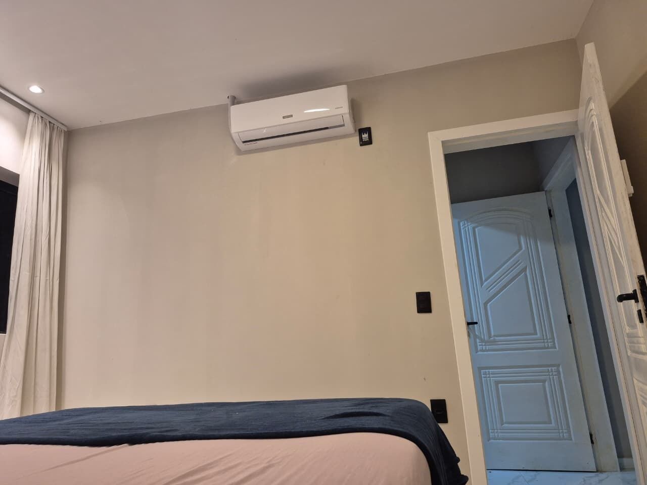 A bedroom with a bed, an air conditioning unit, a door, and a light switch on the wall.