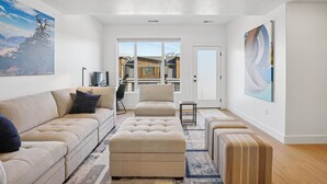 A modern living room with a sofa, ottoman, and a small table. There is a large window with a view of buildings and mountains. - Risen Ranch by Crafty Casitas Hospitality (Mountain Green)