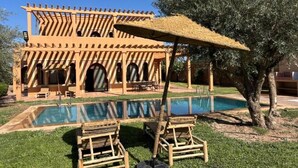Ett område vid poolen med en pergola av trä, solstolar och ett träd.