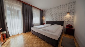 A bedroom with a bed, curtains, a nightstand, a wall-mounted light, and a patterned wallpaper.