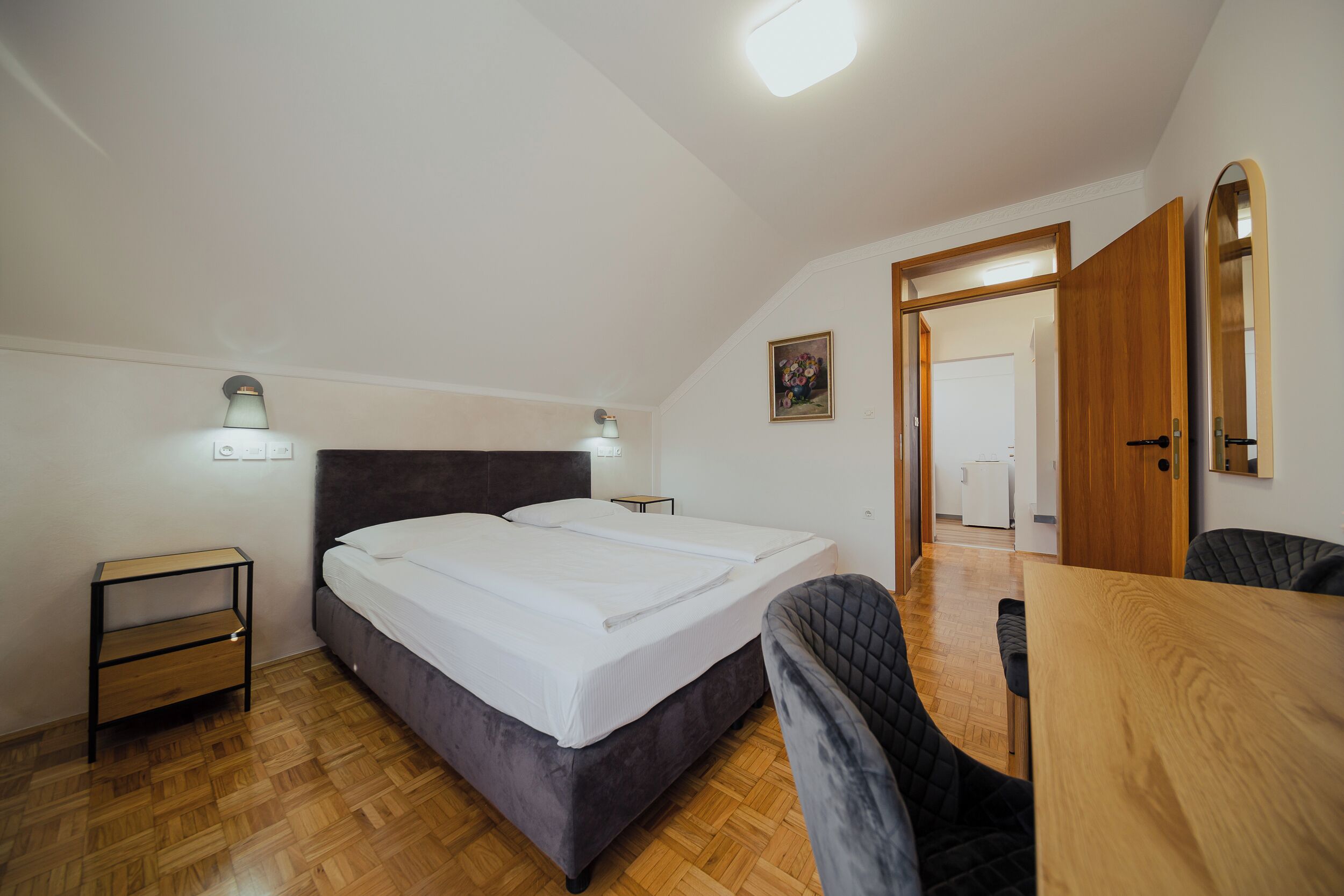 Une chambre à coucher comprenant un lit, des tables de chevet, une chaise, une table en bois et une porte donnant sur une autre pièce.