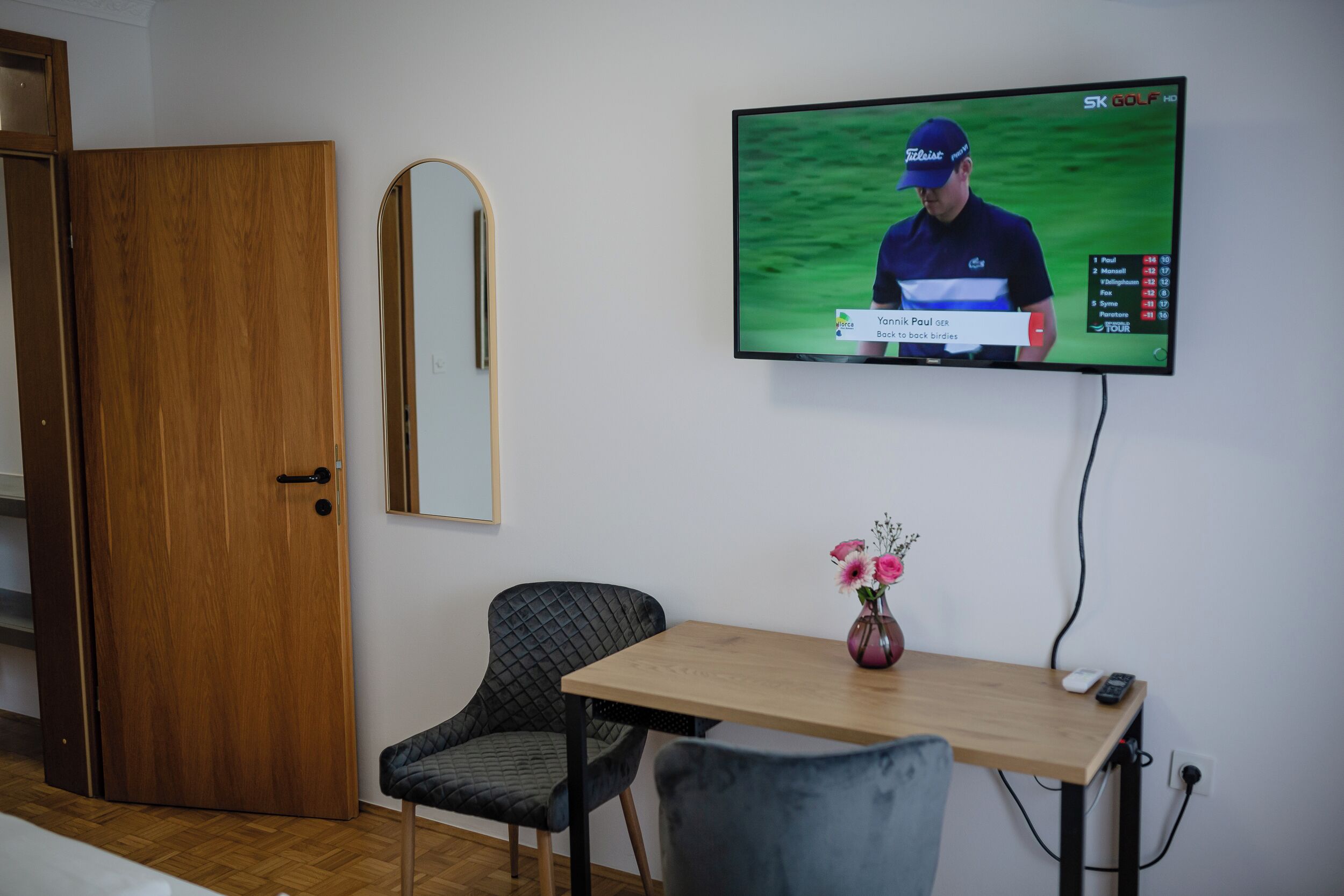 Et værelse med en trädør, et spejl, et TV ophængt på væggen, et lille bord med en vase og en stol.