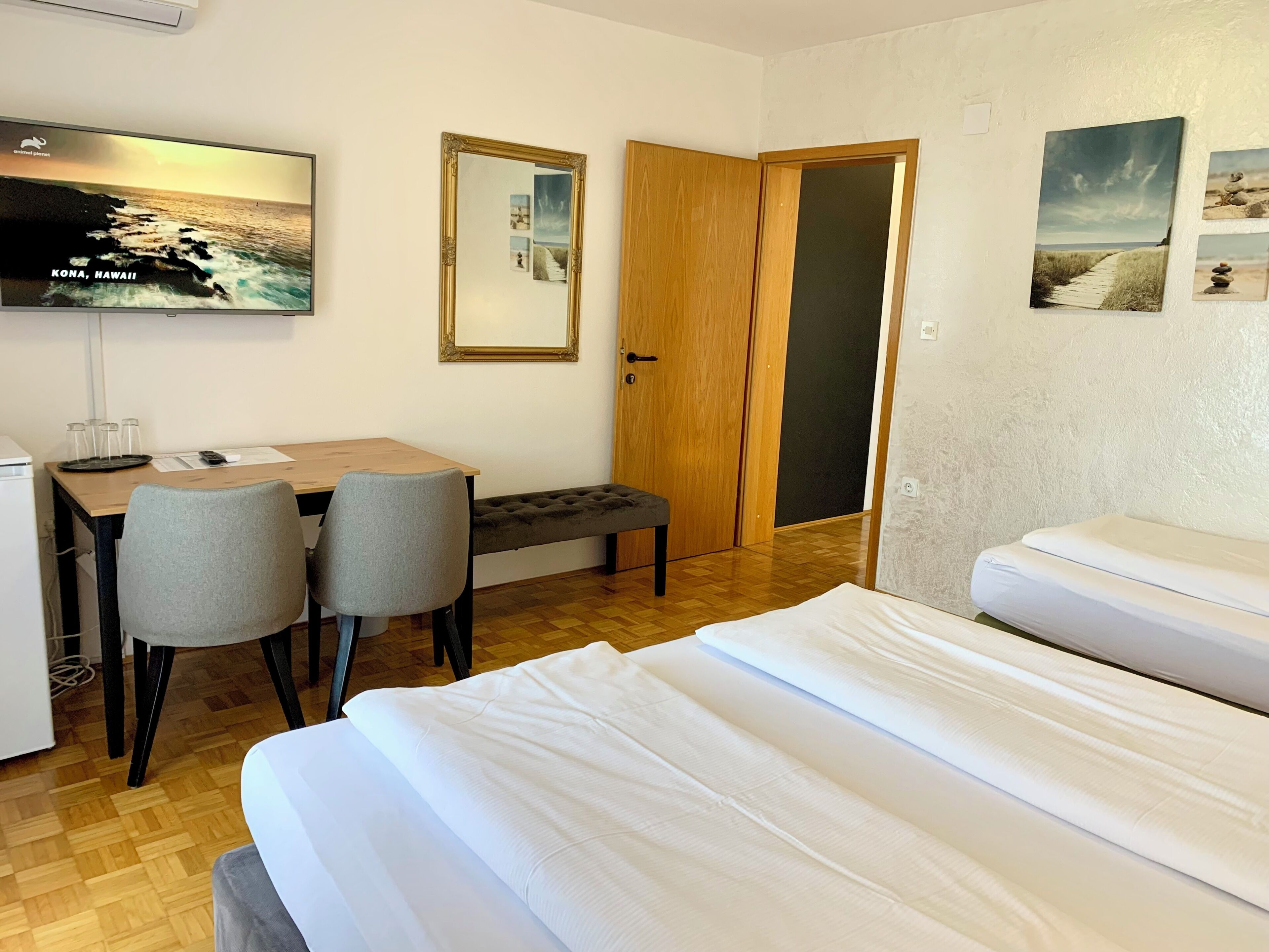 Une chambre d’hôtel comprenant un lit, un bureau avec des chaises, un téléviseur affichant une image de paysage, une porte en bois et un mur orné de tableaux.