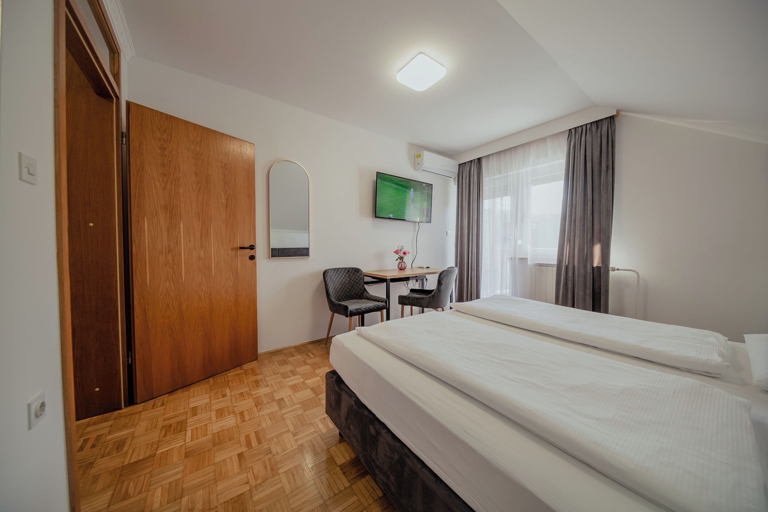 Habitación de hotel con piso de madera, cama, escritorio con silla, televisor y ventana con cortinas.
