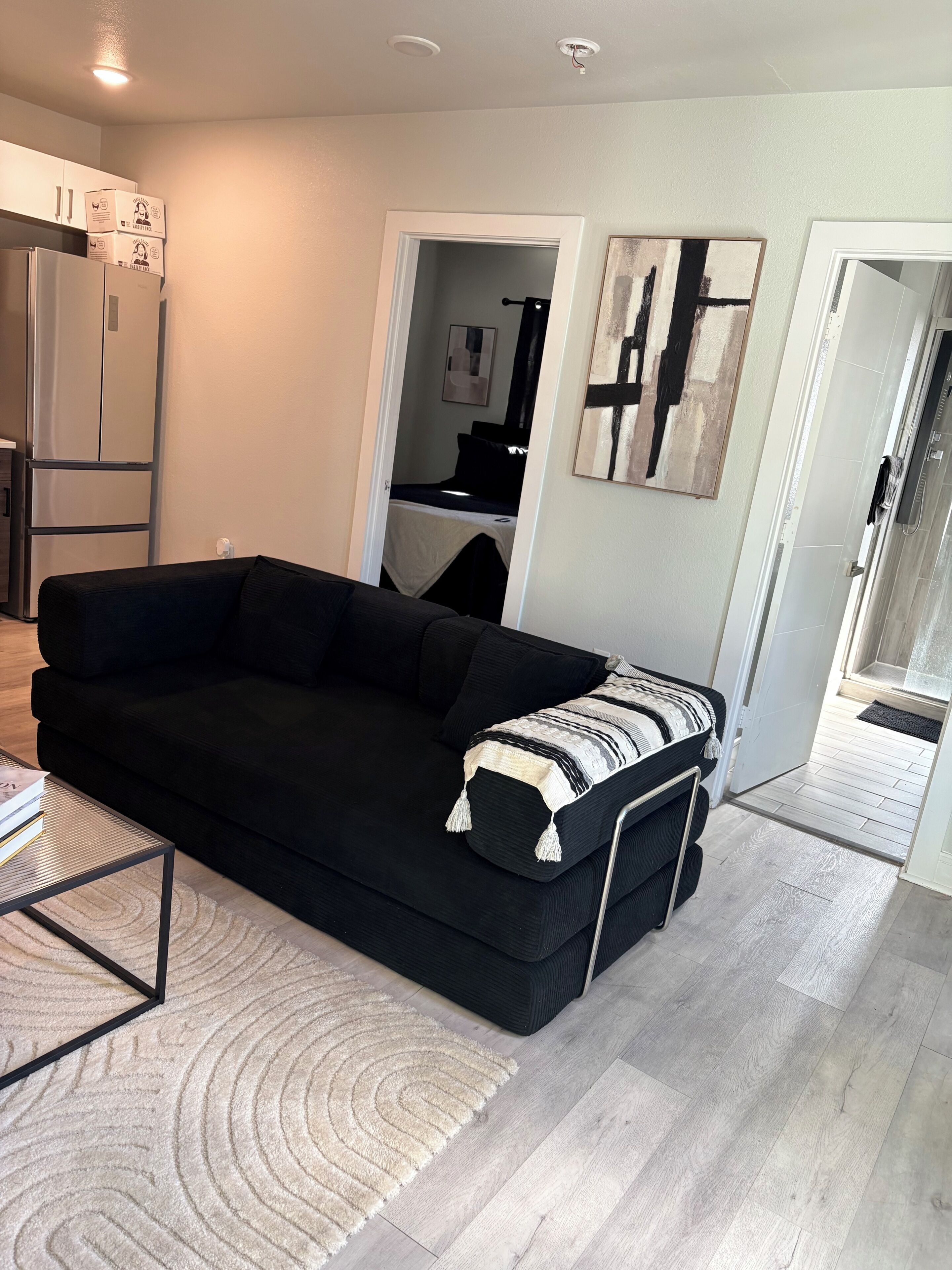A living room with a black sofa, a coffee table, and a rug. There is a kitchen area with a refrigerator and cabinets.