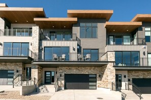 A modern multi-story residential building with a stone facade, large windows, and a covered outdoor area with seating. - Jordanelle Klaim 72 (Hideout)