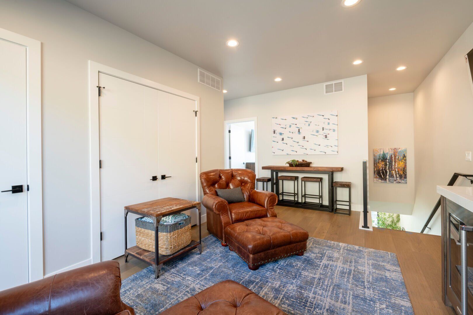 A spacious living area with a brown leather sofa, a small wooden table, and a blue patterned rug. There is a kitchen area with a bar and stools, and a dining table with chairs.