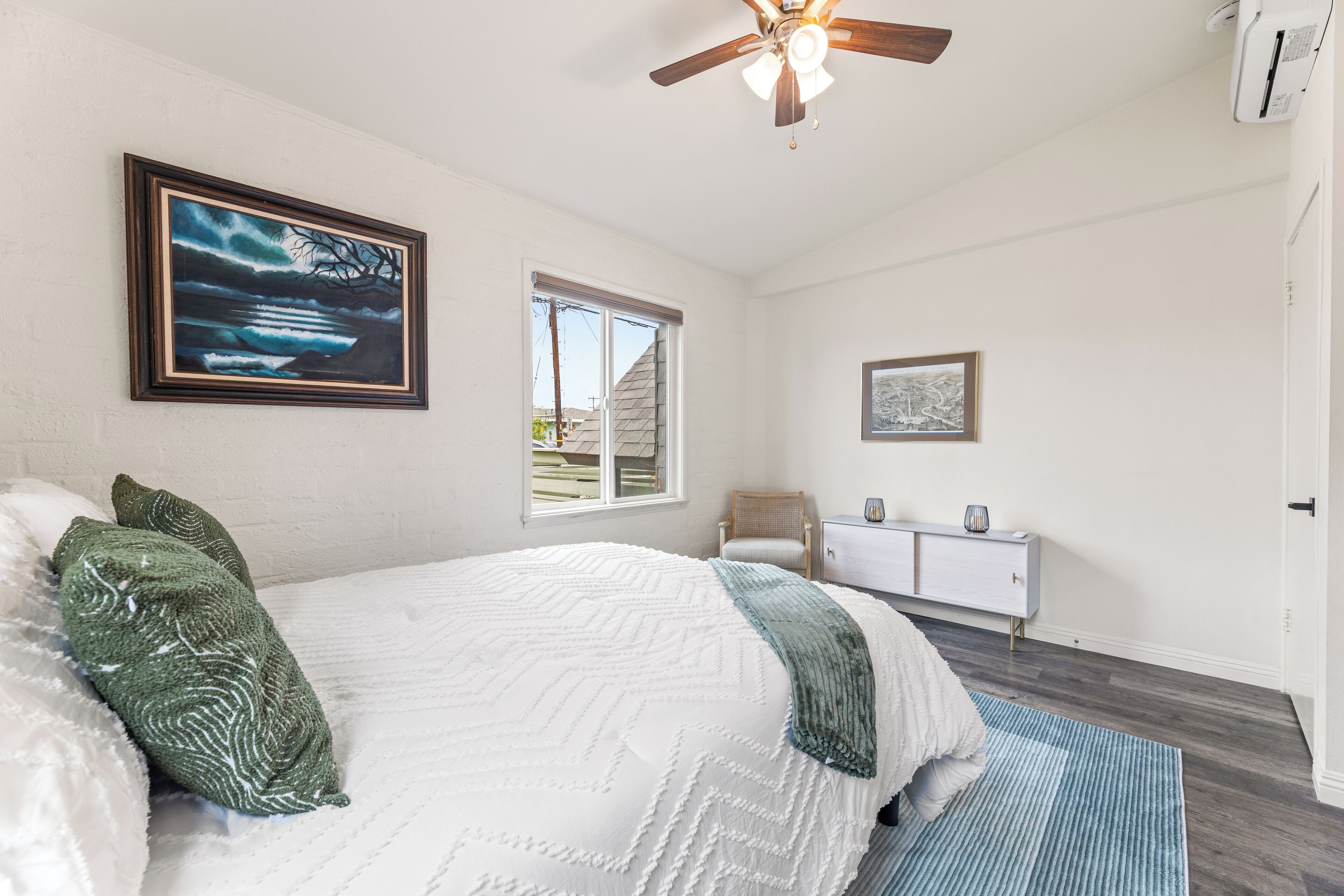 Une chambre avec un grand lit, un ventilateur de plafond, une fenêtre, une chaise, un petit meuble et un tableau accroché au mur.