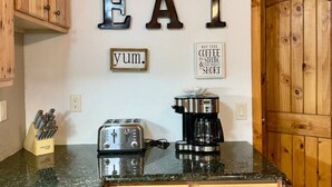 A kitchen with wooden cabinets, a toaster, a coffee maker, and wall decorations including the words "EAT" and "yum." - The Carriage House / Walk to River & Lake, In Town (Grand Lake)