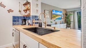 A modern kitchen with a wooden countertop, black sink, and stainless steel appliances. There is a decorative shelf with glassware and a small plant. - Kihei Garden Estates Condo | Pool, Hot Tub, Ground Floor, AC, Walk to Beach (Kihei)