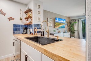 Una cocina moderna con encimera de madera, pileta negra y electrodomésticos de acero inoxidable. Se observa una repisa decorativa con cristalería y una pequeña planta.