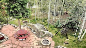 A stone patio with a red metal table and chairs, surrounded by trees and a wooden swing set. - The Red Bears Den / Walk to River & Lake, In Town (Grand Lake)