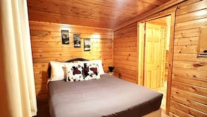 A wooden cabin bedroom with a bed, a nightstand, a wall-mounted light, and a door. - The Red Bears Den / Walk to River & Lake, In Town (Grand Lake)