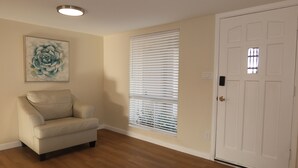 Ein Zimmer mit einer beigen Wand, einer weißen Tür, einem beigen Sessel, einem Fenster mit Jalousien und einer Deckenleuchte.