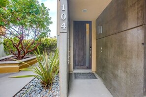 A modern building entrance with a concrete facade, a wooden door, and a small garden area with plants and pebbles. - FULL OCEAN VIEWS AND ROOF TOP DECK IN 3 BR/4 BA OCEANSIDE CONDO / 31 DAY MINIMUM (Oceanside)