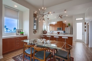 A modern kitchen with wooden cabinets, a glass dining table, and a unique light fixture. - FULL OCEAN VIEWS AND ROOF TOP DECK IN 3 BR/4 BA OCEANSIDE CONDO / 31 DAY MINIMUM (Oceanside)