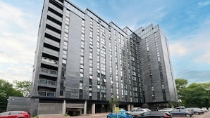 A modern high-rise building with multiple floors, large windows, and a parking area in front. - M16 Urban Green (Manchester)