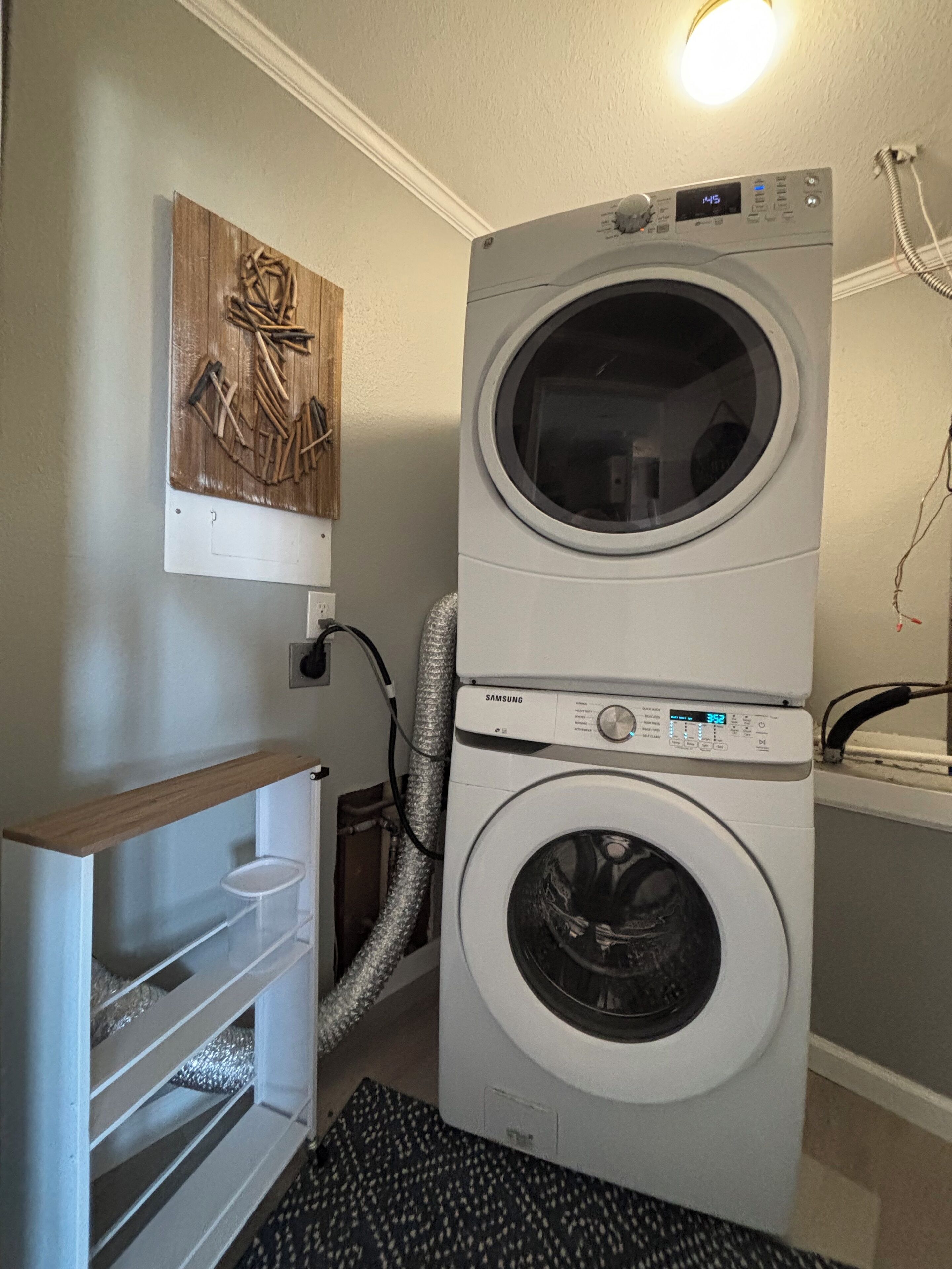 A stacked washer and dryer unit with a control panel, a wooden art piece on the wall, and a white shelf unit with storage baskets.