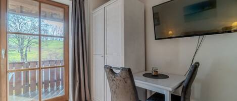 Una habitación con terraza de madera, puerta corrediza de vidrio, una mesa blanca con dos sillas, televisor montado en la pared y una pequeña vela sobre la mesa.