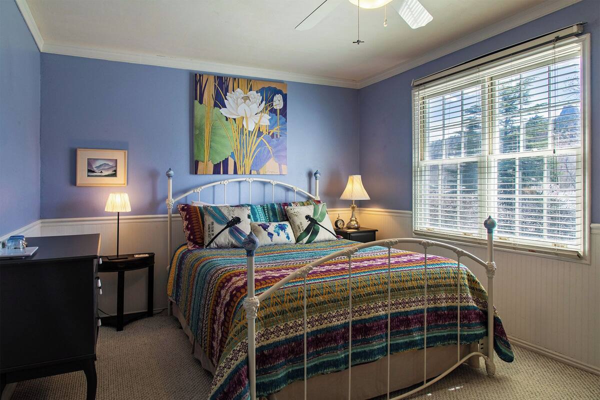 A bedroom with a bed, two bedside tables, a desk, a window with blinds, and a painting on the wall.