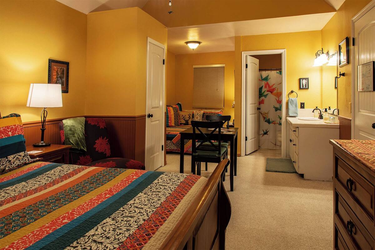A bedroom with a bed, a desk, a chair, a dresser, and a bathroom with a sink and mirror.