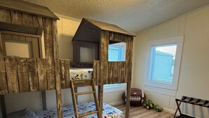 A room with a bunk bed, a ladder, a small playhouse, and a window with blinds.