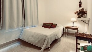 Een keurig opgemaakte slaapkamer met een bed, een nachtkastje met een lamp en een wandplank met decoratieve objecten.