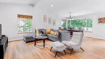 A spacious living room with a sectional sofa, a round coffee table, and a grey armchair. There are large windows providing natural light and a view of greenery outside.