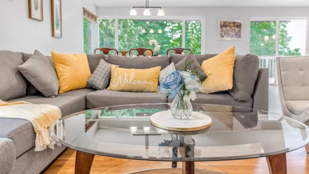 A modern living room with a glass coffee table, grey sofa with yellow cushions, and a 'Welcome' pillow. - Spacious 6BR/4BA | Close to Airport @South Seattle (Tukwila)