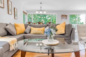 A living room with a grey sofa, yellow cushions, a glass coffee table with a vase of flowers, and a 'Welcome' pillow. - Spacious 6BR/4BA | Close to Airport @South Seattle (Tukwila)