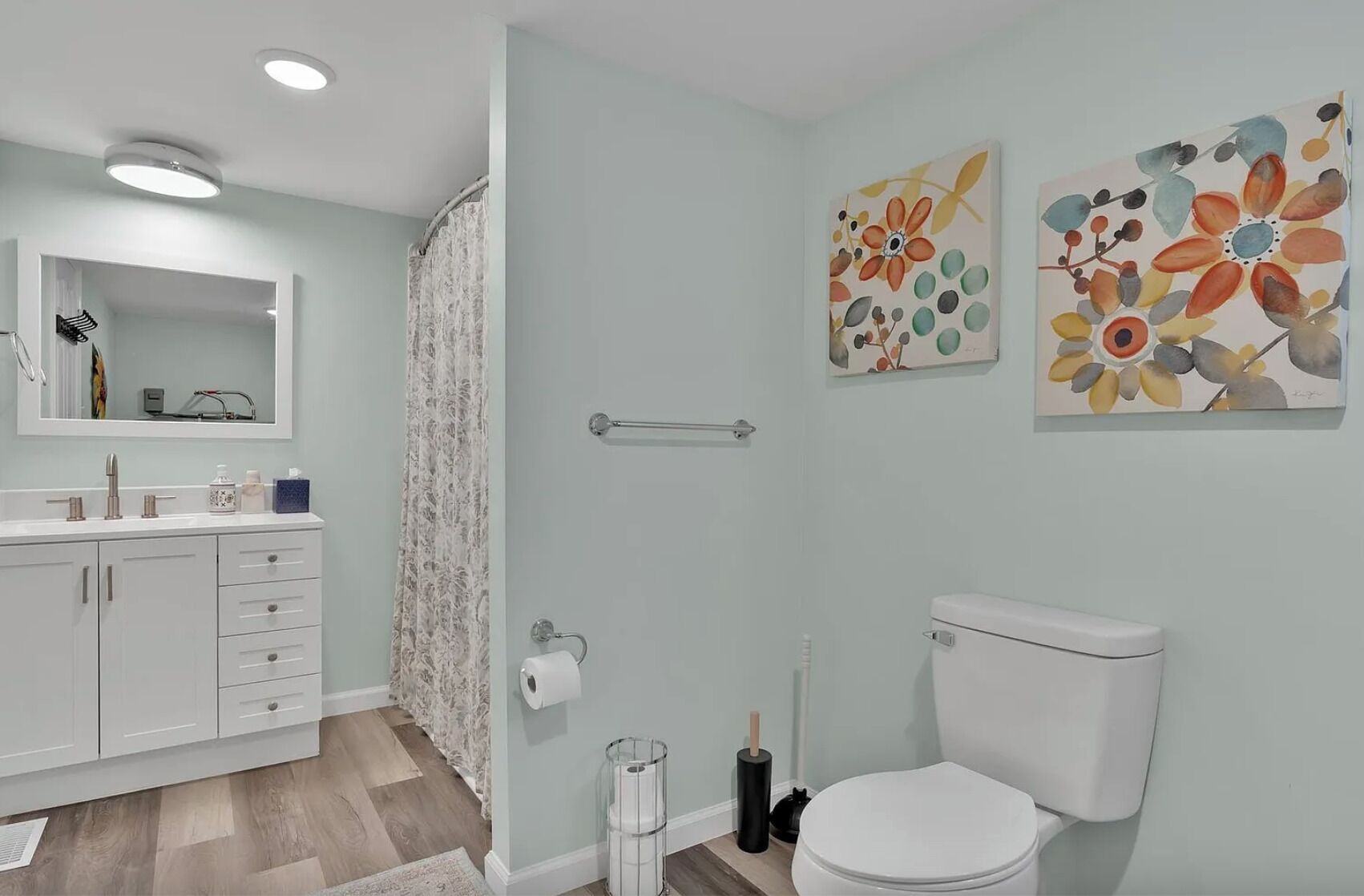 A bathroom with a toilet, sink, and shower. There are two floral paintings on the wall.