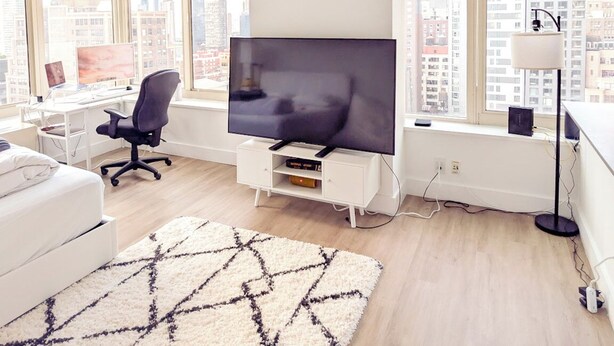 Un soggiorno moderno con una televisione a schermo piatto, un divano bianco, una sedia da ufficio nera, un tappeto decorato e ampie finestre con vista sulla città.