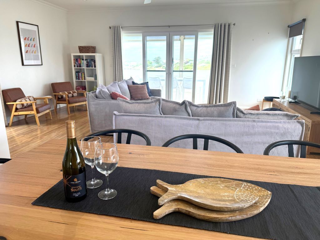 Una sala de estar con sofá, sillas, una mesa de comedor, una botella de vino y copas, y un amplio ventanal con vistas.