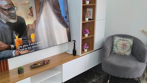 A modern living room with a flat-screen TV, a wooden shelf with decorative items, a grey armchair, and a marble floor.