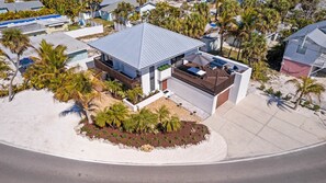 Una casa moderna con un porche cubierto, rodeada de palmeras y un paisaje arenoso.