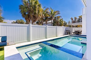 Ein Schwimmbad mit weißem Zaun, eine Terrassenanlage mit Gartenmöbeln und Palmen im Hintergrund.
