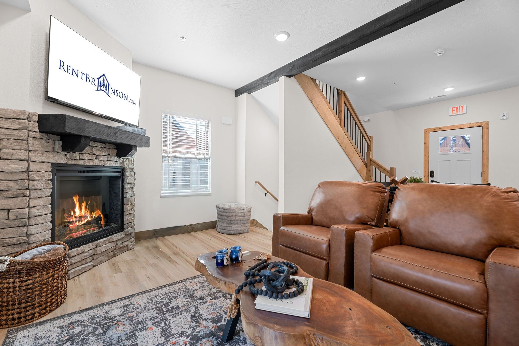 A living room with a stone fireplace, leather sofa, and a television displaying 'RENTBRINSON.COM'.