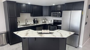 A modern kitchen with dark cabinets, a central island, stainless steel appliances, and a marble countertop. - The Pacibeach Condo - Oceanfront • Infinity View (Playas de Rosarito)