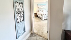 Een gang die leidt naar een slaapkamer met een bed, een nachtkastje en een kunstwerk aan de muur.