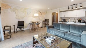 Ein modernes Wohnzimmer mit einer großen Weltkarte an der Wand, einem grauen Sofa, einem Couchtisch mit Zeitschriften und einem Essbereich mit kleinem Tisch und Stühlen.