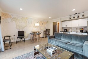 A modern living room with a large world map on the wall, a grey sofa, a coffee table with magazines, and a dining area with a small table and chairs. - Wall Street Wander - Steps to Downtown & River (Bend)