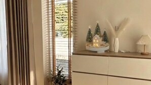 Ein modernes Interieur mit Holzboden, einem Glastisch, einer Vase mit Blumen, einem weißen Schrank mit Schubladen und einem Fenster mit Jalousien.