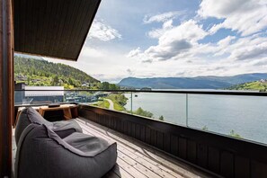 Un balcon doté d’un plancher en bois et d’un canapé gris, offrant une vue sur un lac et des collines lointaines.